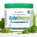 A white plastic tub of Allmax CytoGreens Premium Green Superfood with a bright green lid, surrounded by fresh green spinach leaves and a pile of green powder on a white background.